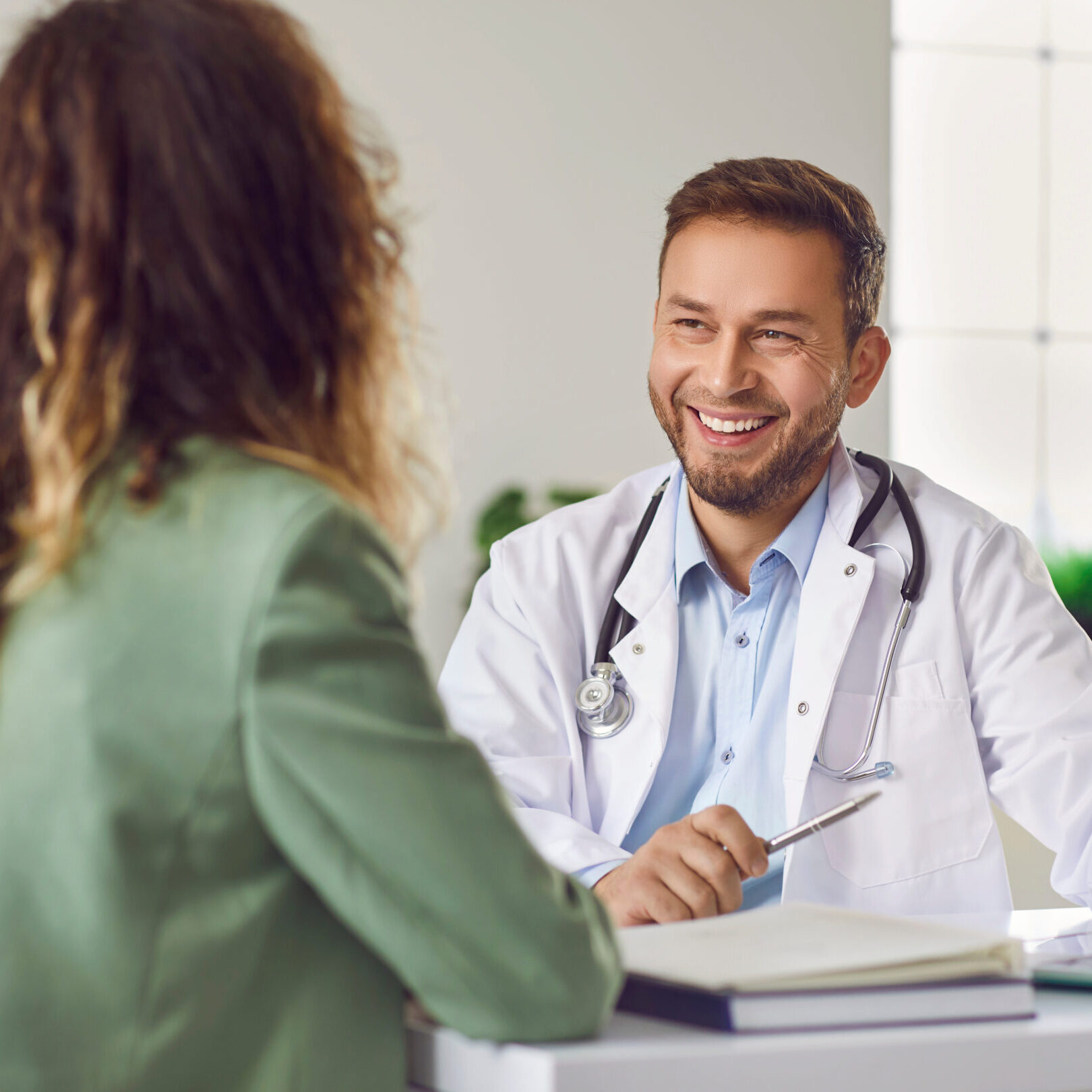 Woman visiting a doctor at the medical office during medical examination. Cheerful friendly male doctor at medical office in hospital, showing examination results on laptop monitor screen and having consultation with young woman patient sitting in clinic.